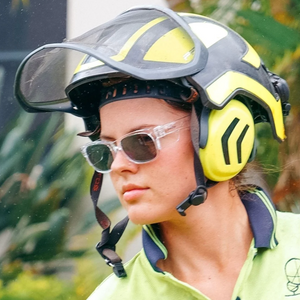 A person wearing SafeStyle Fusions Clear Frame Polarised Lens safety sunglasses, a green and black protective helmet with a visor and ear protection, and a green shirt is outdoors with blurred greenery in the background, enjoying effective UV protection.