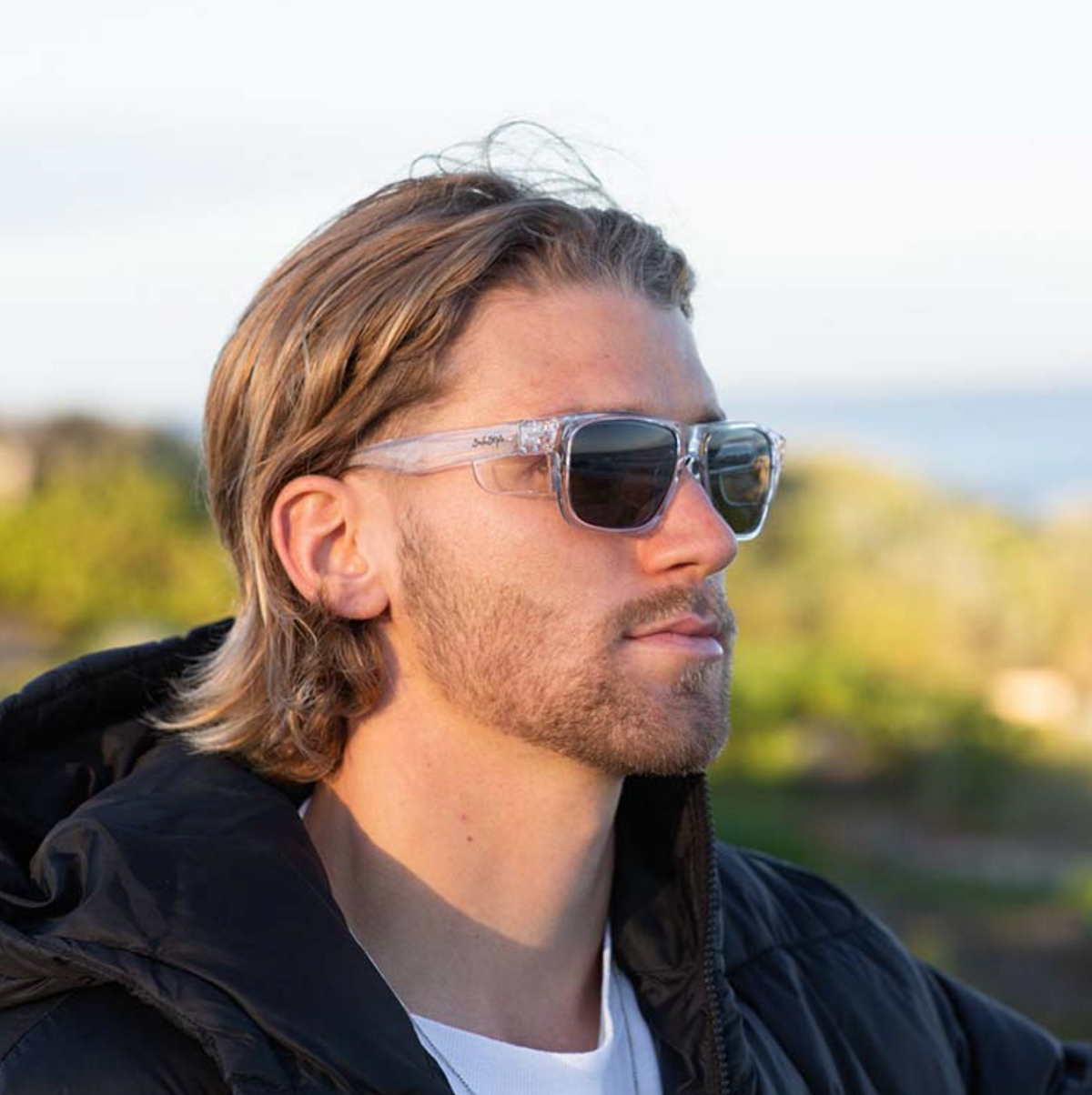 A man with shoulder-length blond hair and a beard wears SafeStyle Fusions Clear Frame Polarised Lens sunglasses with UV protection, paired with a black jacket, standing outdoors against a blurred green and blue background.