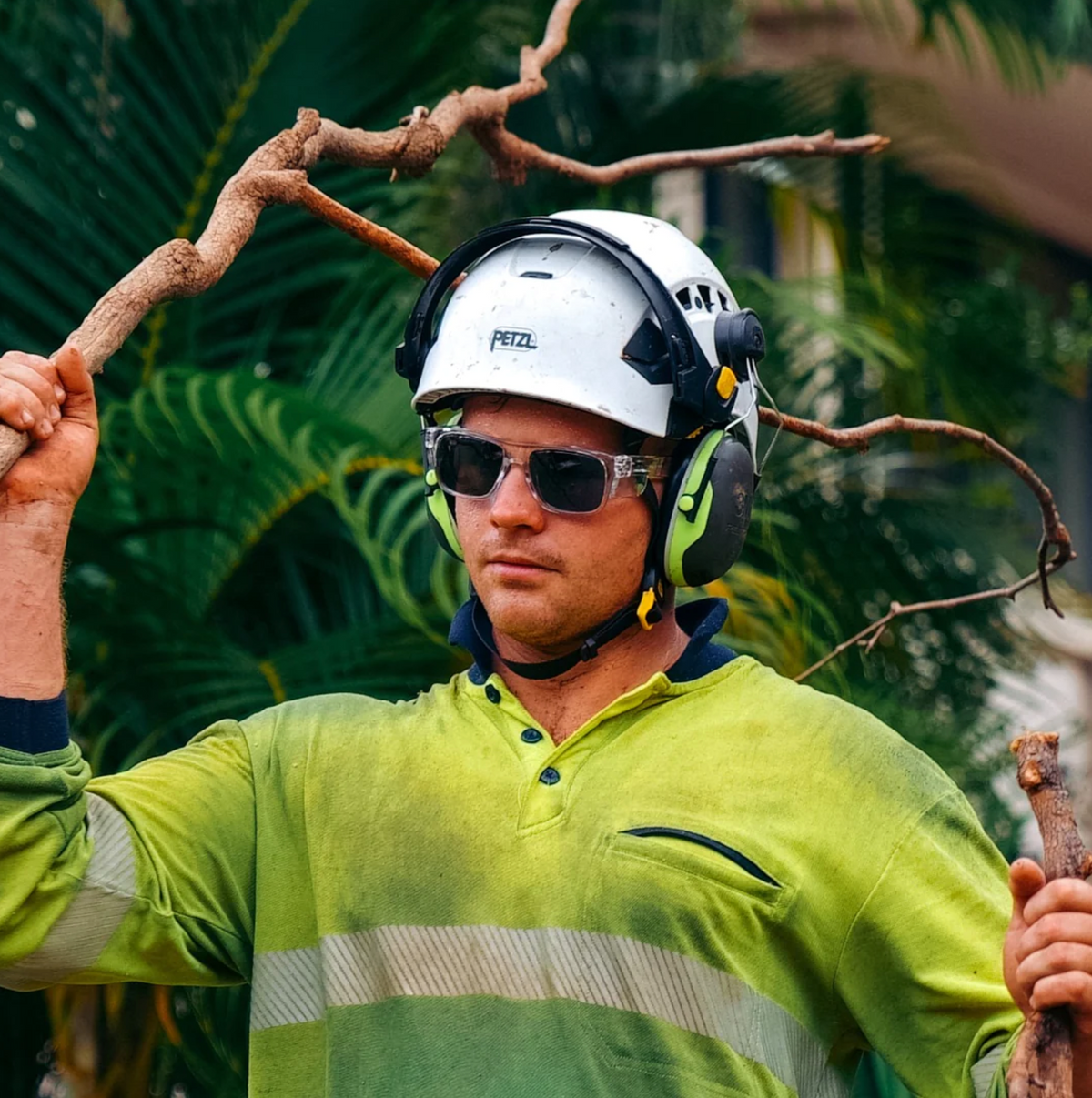 Wearing a helmet and SafeStyle Fusions Clear Frame Tinted Lens glasses with UV400 protection, and earmuffs, a man holds a tree branch on his shoulder. He is in a green high-visibility shirt, standing before lush greenery.