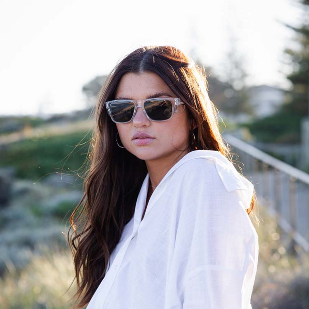 A woman with long hair wears SafeStyle Fusions Clear Frame Tinted Lens sunglasses and a white shirt outdoors. The background is blurred with greenery and a fence, hinting at nature. Bright lighting suggests it's a sunny day ideal for UV400 protection.