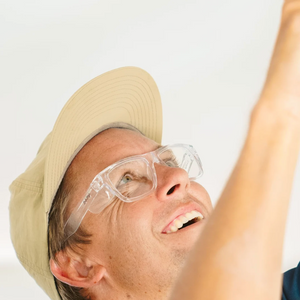 A person in a beige cap and SafeStyle Fusions Clear Frame Clear Lens safety glasses smiles as they look upwards against a plain light background, highlighting their expression and pose.