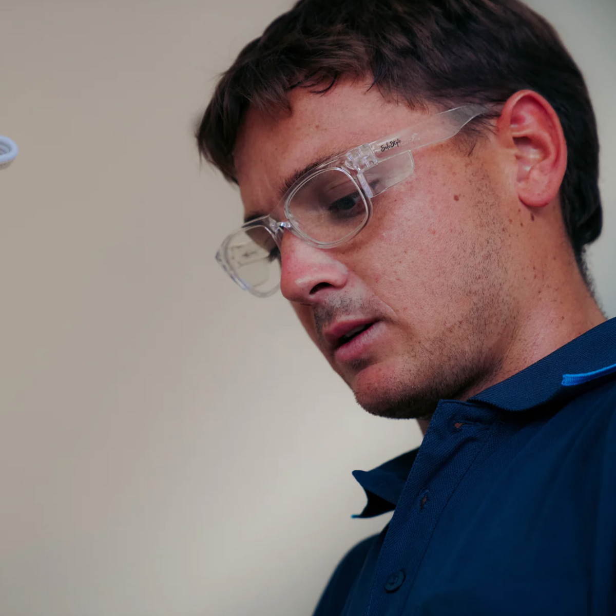 A man wearing SafeStyle Classics Clear Frame Clear Lens safety glasses and a navy blue collared shirt looks down thoughtfully against a plain background.