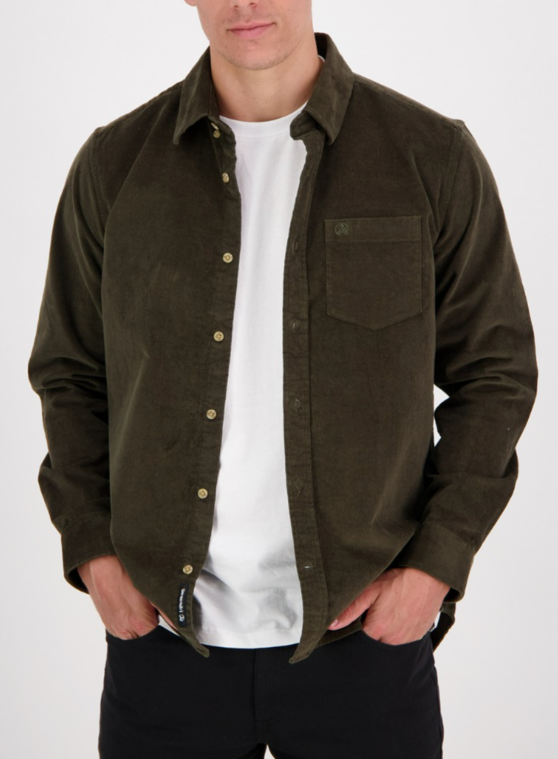A man in a Swanndri Ranfurly Corduroy Shirt, dark olive green and button-up, stands with hands in pockets against a plain background over a white t-shirt, showcasing a tailored fit that exudes effortless style.