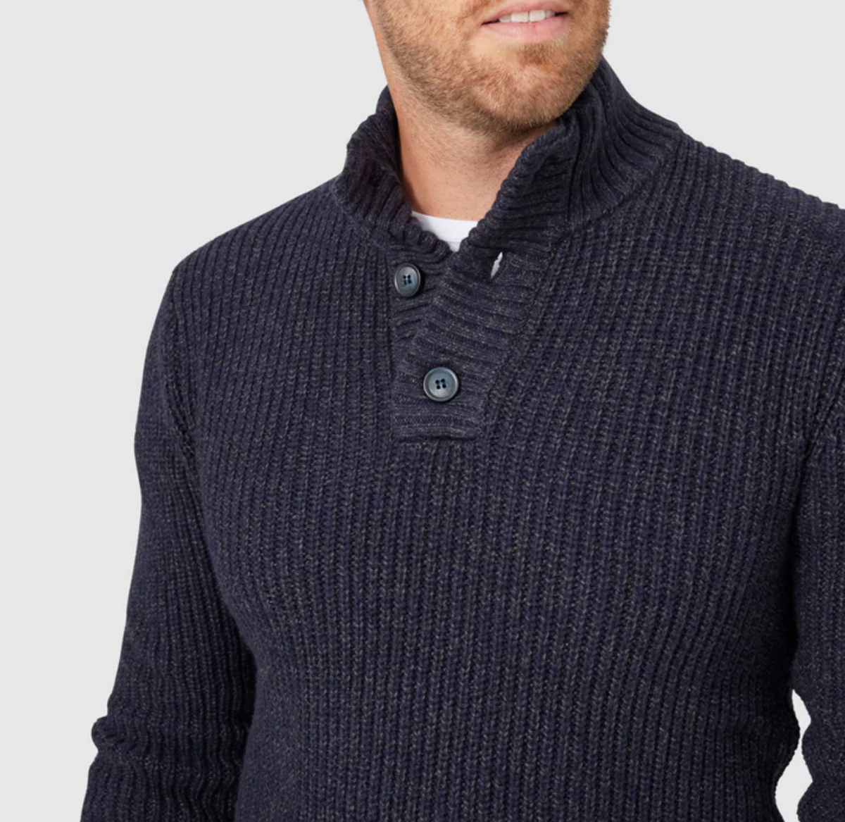 A man models the Blazer Oscar Button Neck Knit—a dark blue ribbed sweater featuring a two-button neck design, ribbed cuffs, and waistband—against a light gray background. Only his upper torso and part of his face are visible.