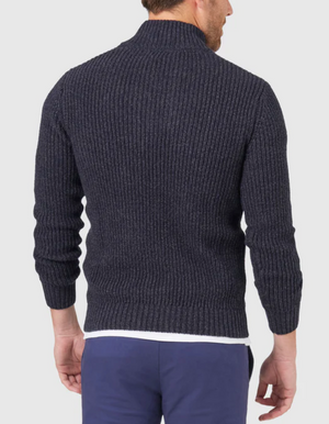 A man in a dark grey Blazer Oscar Button Neck Knit sweater with ribbed cuffs and waist faces away from the camera. His short hair complements his blue pants, and a white shirt subtly peeks out beneath against the plain light grey background.