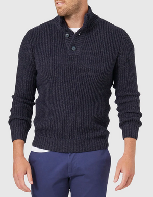 Wearing the Blazer Oscar Button Neck Knit, a dark ribbed sweater with ribbed cuffs and waist, paired with dark blue pants, the person stands out against the plain light background. Their head is slightly turned, concealing their features.