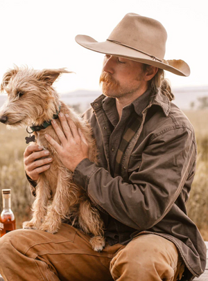 A person outdoors in a cowboy hat and an Outback Loxton Jacket with polar fleece lining sits with a scruffy dog. A bottle is on the ground beside them amidst a blurred landscape of fields and hills.