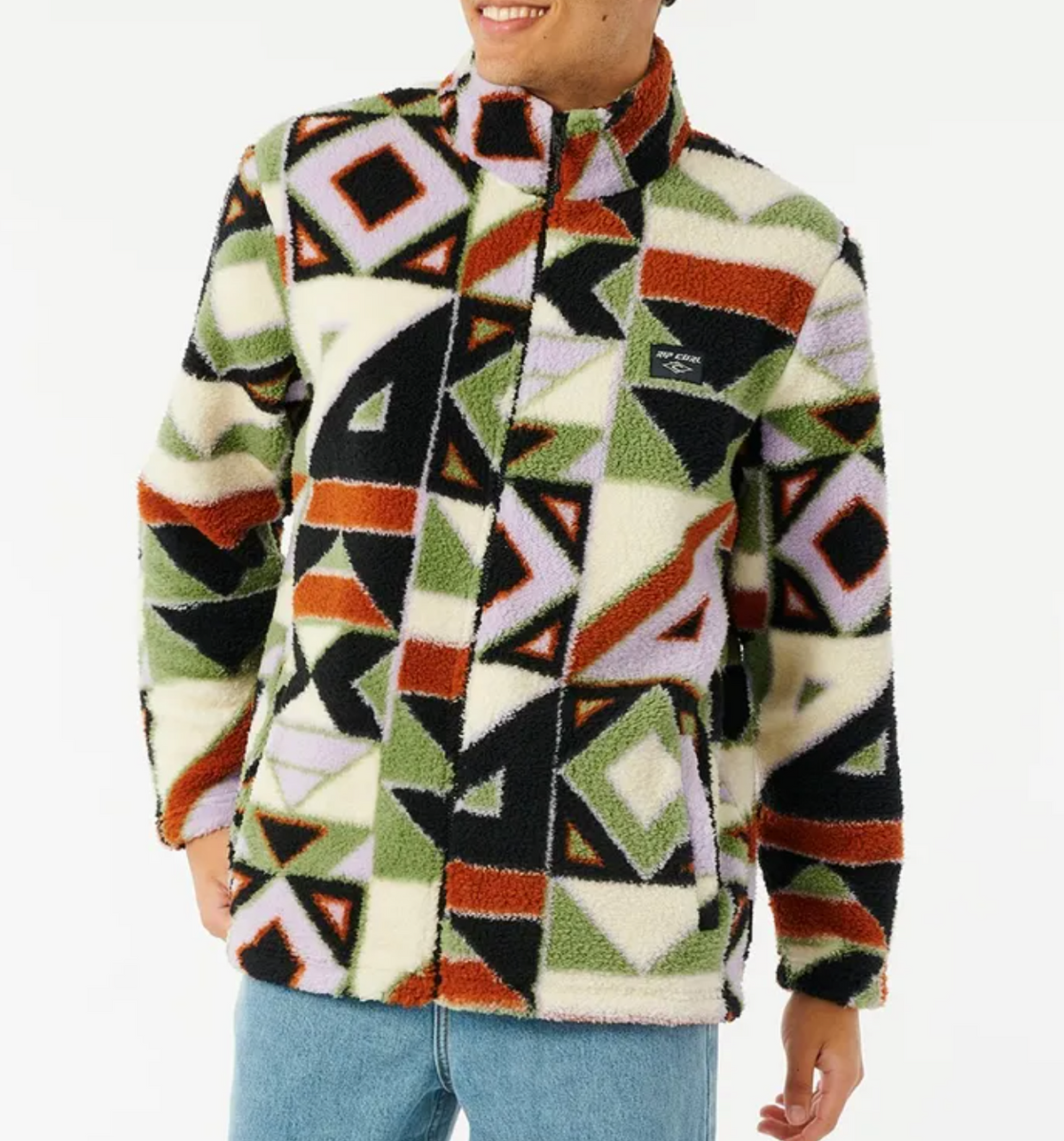 A person wears the Rip Curl Nah Yeh Polar Fleece, a fuzzy zip-up in bold black, olive green, burnt orange, lavender, and cream geometric patterns, paired with light blue jeans, smiling against a plain white background.