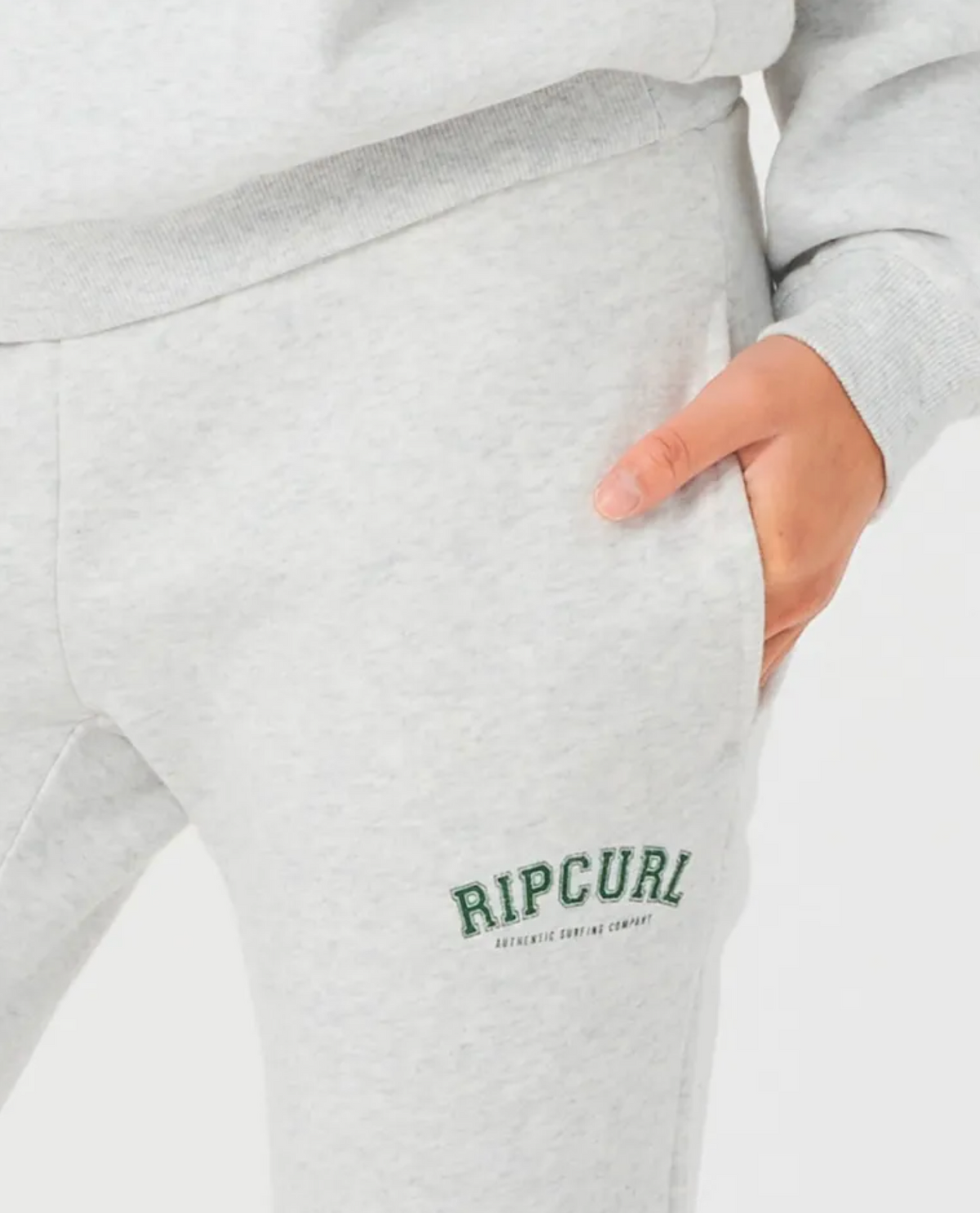 Close-up of someone wearing Rip Curl Varsity Pants in light gray with a hand in the pocket. "RIPCURL" is embroidered in green on the left side, and they have a cozy fleece lining. The background is bright white.