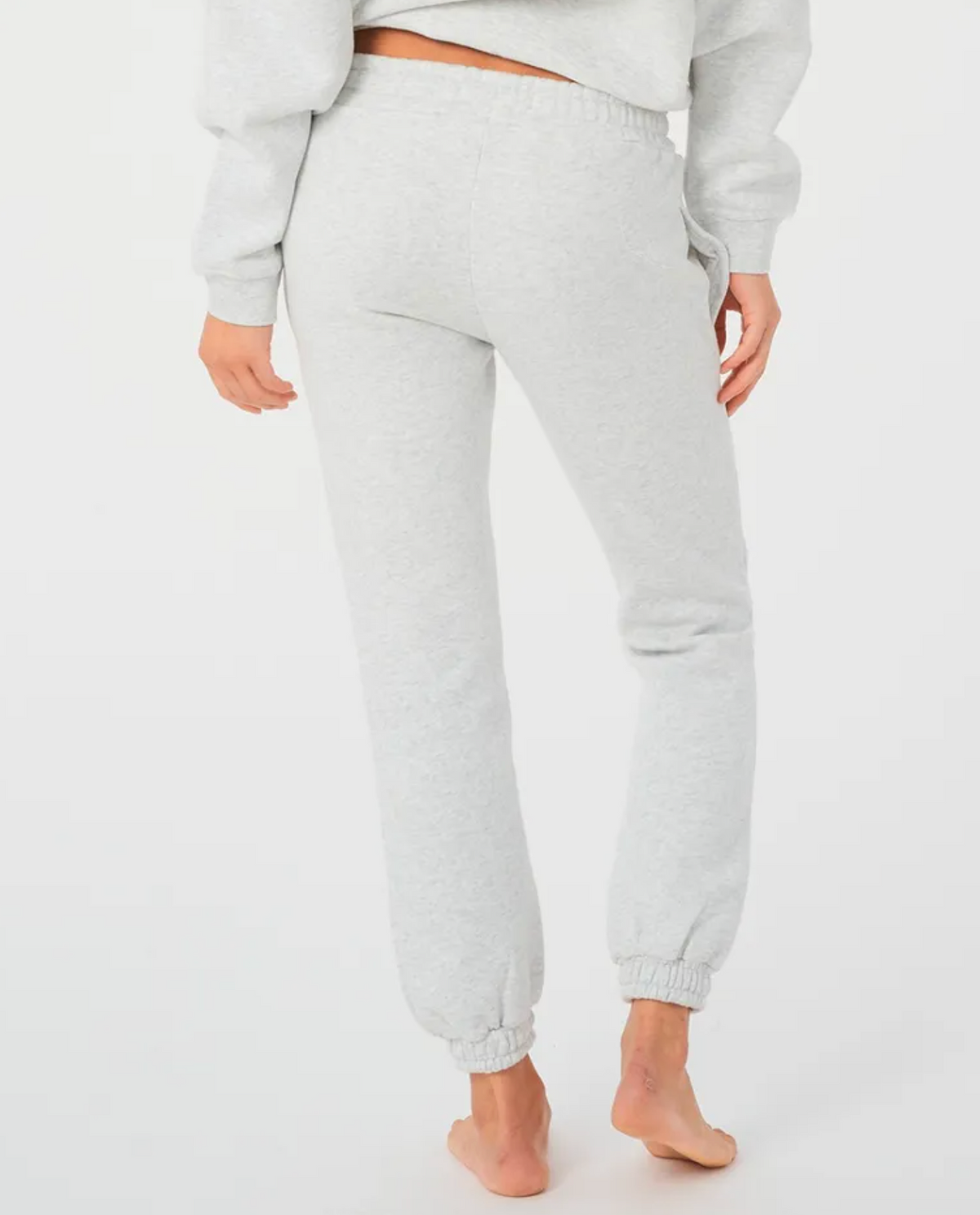 Standing barefoot on a white background, the person wears light gray fleece-lined Rip Curl Varsity Pants with elasticated cuffs and a matching sweatshirt, facing away and placing their right hand in their pocket.