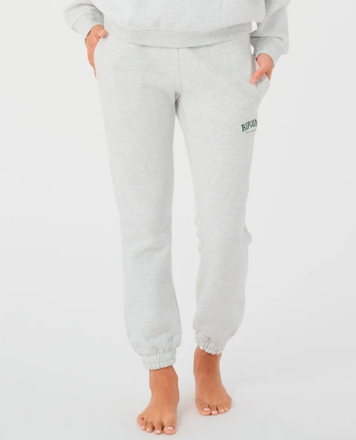 A person stands barefoot against a white background, wearing Rip Curl Varsity Pants in light gray with "RIP2023" in green on one side. With an elastic waistband and cuffs, these pants feature cozy fleece lining. A matching light gray top is partially visible.