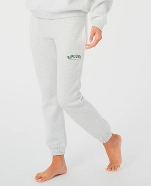 A person wearing Rip Curl Varsity Pants, featuring "Rip Curl" in green on the left leg, stands barefoot on one foot with the other slightly bent. The light gray trackpants offer comfort with a fleece lining against the plain white background.