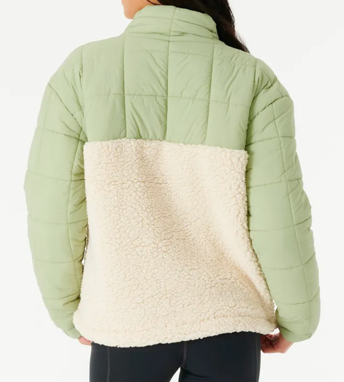 A person is wearing the Rip Curl Anti Series Anoeta Fleece, featuring light green quilted fabric on the shoulders and sleeves, cream-colored sherpa fleece on the lower back, and a relaxed fit. They face away with long dark hair, paired with black pants.