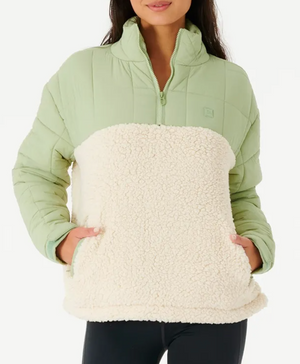 A woman wears the Rip Curl Anti Series Anoeta Fleece, featuring a relaxed fit with a light green quilted top and cream fleece bottom. Its half-zip design and side pockets make it perfect to pair with her black pants.