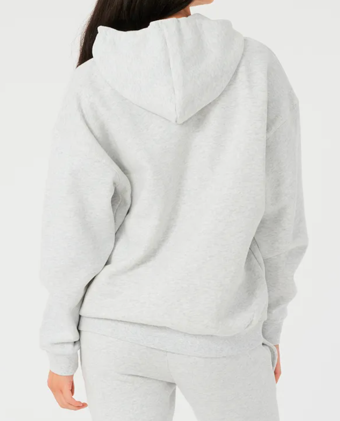 A person with long dark hair faces away, wearing a light gray Rip Curl Varsity Hoodie and matching sweatpants. The relaxed fit ensures comfort, with the hood down revealing the soft fleece lining against a plain white background.