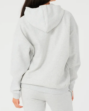 A person with long dark hair faces away, wearing a light gray Rip Curl Varsity Hoodie and matching sweatpants. The relaxed fit ensures comfort, with the hood down revealing the soft fleece lining against a plain white background.