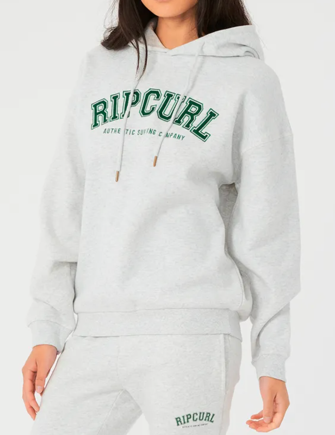 A person is dressed in a comfortable and casual Rip Curl Varsity Hoodie, which is fleece-lined with a gray relax fit and drawstring. "Ripcurl" is printed in green on both the chest of the hoodie and matching sweatpants.