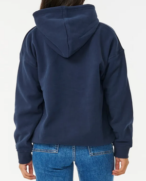 Rear view of a person in a cozy, dark blue Rip Curl Varsity Hoodie with the hood up and paired with blue jeans against a crisp white background.