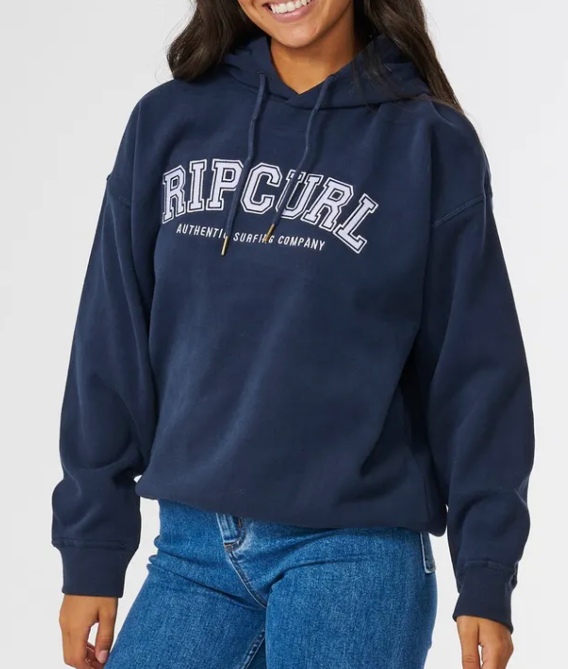 A person wearing a Rip Curl Varsity Hoodie with "RIP CURL AUTHENTIC SURFING COMPANY" printed on the front, paired with blue jeans, smiles casually while standing against a plain background.