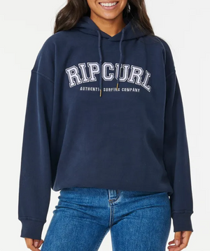 Someone is wearing a navy blue Rip Curl Varsity Hoodie, known for its authentic surfing vibe and comfy fit, featuring "Authentic Surfing Company" in white. It's paired with blue jeans against a plain white background for a casual look.