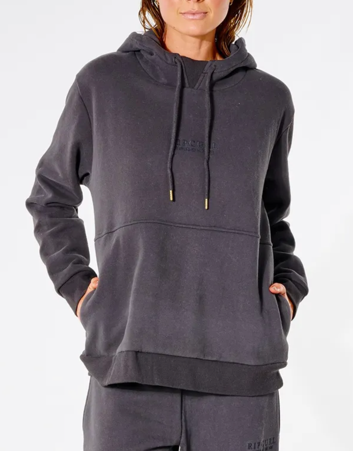A person is wearing a comfortable Rip Curl Premium Surf Hoodie and matching dark grey sweatpants. The relaxed fit hoodie has drawstrings, with the person's hands in its front pockets, set against a plain white background.