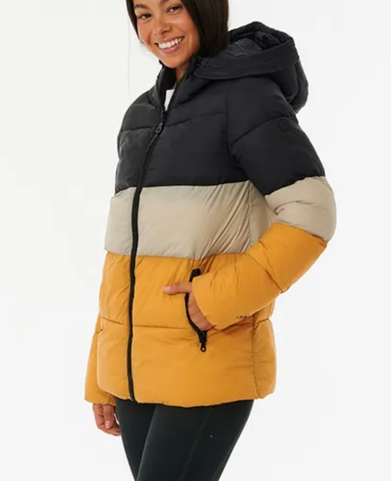 A smiling person wears the Rip Curl Anti Series Insulated II Jacket, showcasing bold black, beige, and mustard yellow blocks. This hooded puffer features DWR water-repellent tech, a front zipper, and pockets. Styled with black pants against a plain white background.