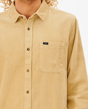 A person in a Rip Curl State Cord Shirt, a beige cotton corduroy long-sleeve with a front pocket and dark buttons, stands against a white background, flaunting their curly hair.