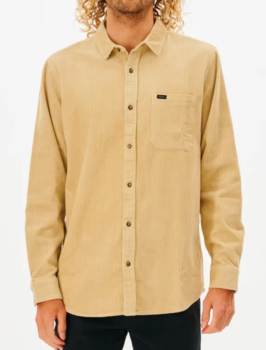 A person with curly hair sports the Rip Curl State Cord Shirt in beige, a long-sleeve corduroy design featuring a chest pocket and a subtle logo tag, paired with black pants. The cotton shirt contrasts against the plain white background.