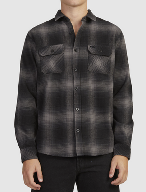 A person is wearing a Rip Curl Count Flannel Shirt—a black and gray plaid button-up with long sleeves and two chest pockets—made from soft brushed cotton, paired with black pants against a plain white background.