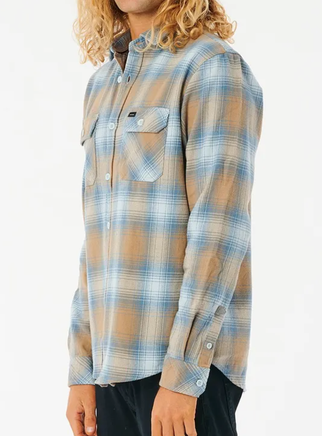 A person with curly blonde hair wears a Rip Curl Count Flannel Shirt in blue and brown plaid, made of brushed cotton with buttoned chest pockets. It's buttoned up and paired with dark pants as they stand against a plain white background.