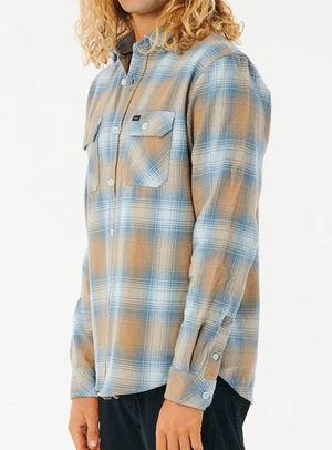 A person with curly blonde hair wears a Rip Curl Count Flannel Shirt in blue and brown plaid, made of brushed cotton with buttoned chest pockets. It's buttoned up and paired with dark pants as they stand against a plain white background.