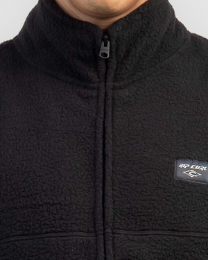 Someone wears the Rip Curl Rincon Zip Crew Polar Fleece, a black textured polyester jacket with a high collar and visible vertical zipper. The chest features a small rectangular label with the Rip Curl logo. The background is plain light gray.