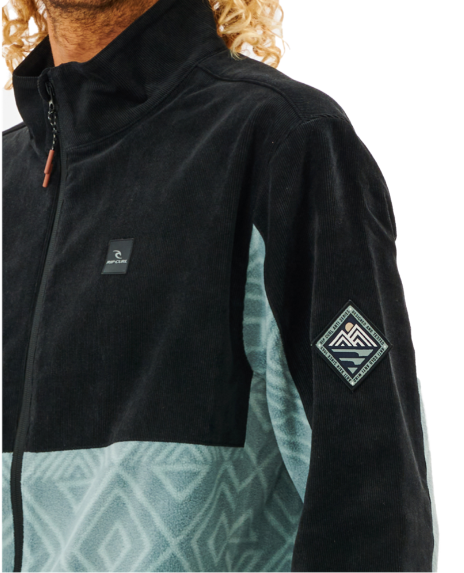 A person with curly hair is wearing a zipped Rip Curl Anti Series Journey Fleece jacket. It features a black upper section, patterned light blue lower section, and geometric mountain motif patches on the chest and sleeve. The water-repellent design includes zippered pockets.