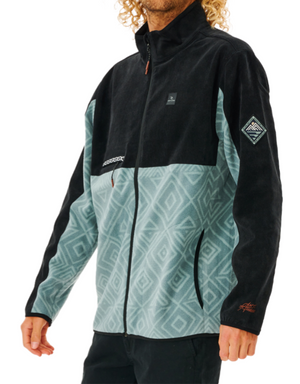 The man wears a Rip Curl Anti Series Journey Fleece with black upper sections and patterned light teal lower sections. This water-repellent fleece includes a full zip, long sleeves, and logos on the chest and left sleeve. Both stylish and functional, it complements his curly hair perfectly.
