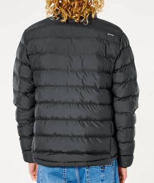 A person with curly hair is wearing a Rip Curl Anti Series Elite Puff Crew Jacket in black, filled with premium duck down, and blue jeans, facing away from the camera.