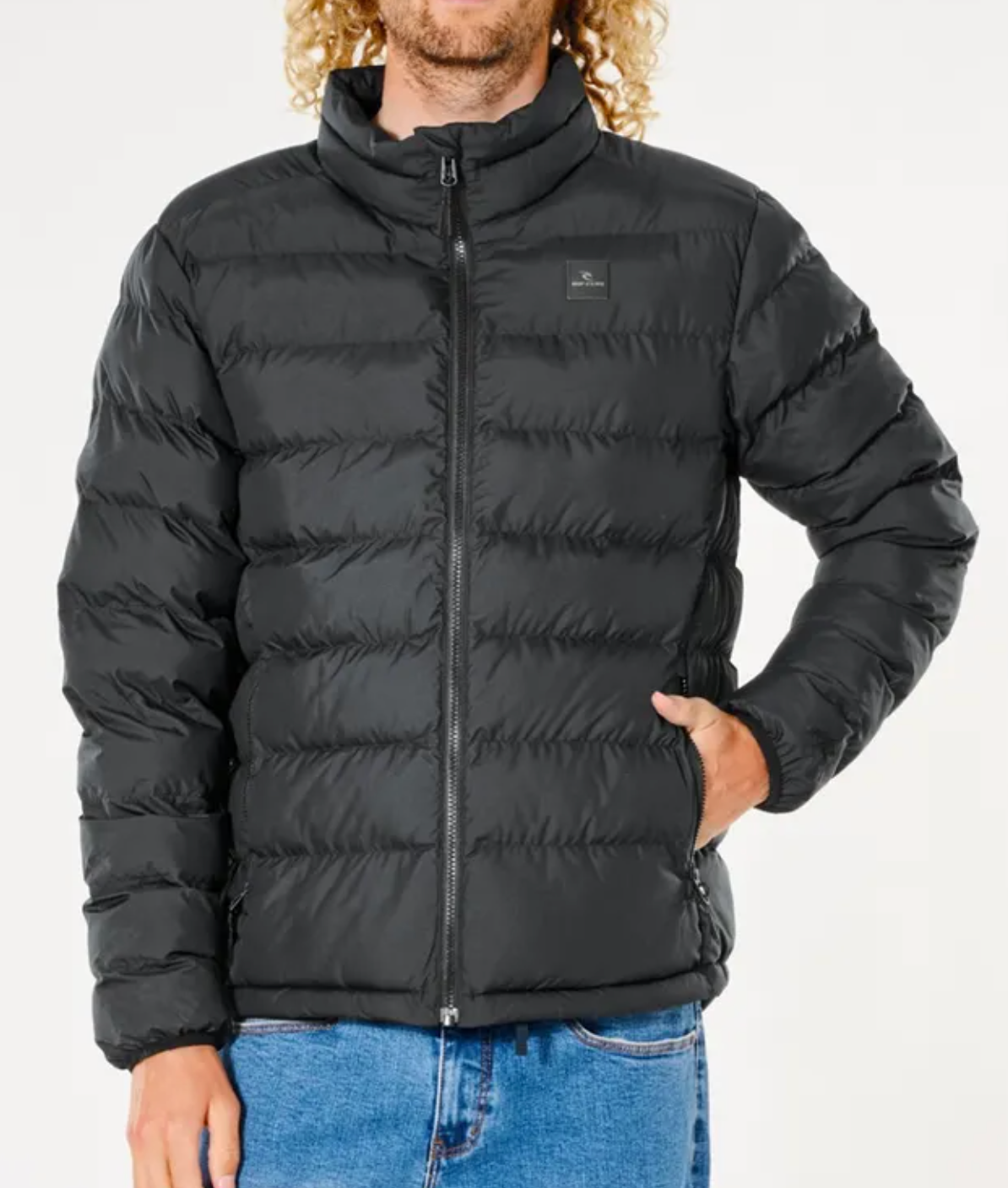 A person with curly hair wears a black Rip Curl Anti Series Elite Puff Crew Jacket made from premium duck down and blue jeans, standing against a plain white background. One hand is in the jacket pocket, which features a small square logo on the chest.