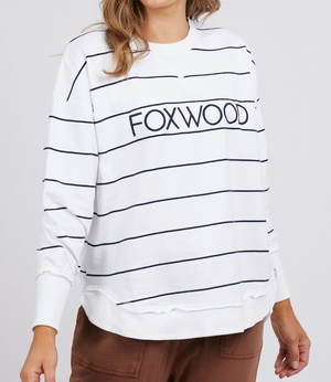 The person wears the Foxwood Simplified Stripe Crew—a relaxed fit white sweatshirt with black stripes and "FOXWOOD" lettering, crafted from 100% cotton. It's paired with brown pants for a stylishly comfortable look.