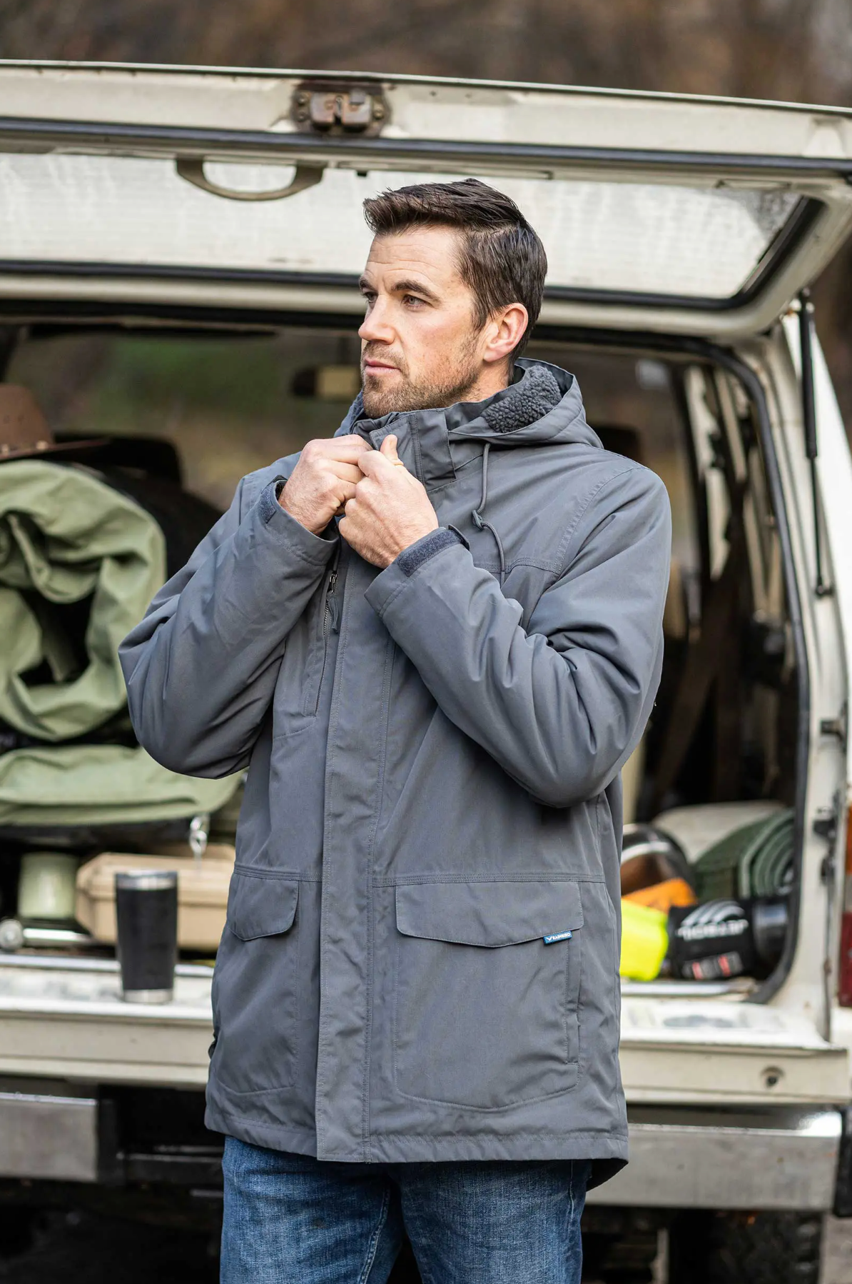 A man in a gray Rainbird Altair Jacket adjusts his hood near an open SUV filled with camping gear. Confident in his windproof and waterproof jacket's fleece lining, he looks ready for outdoor activities.