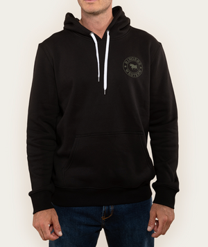 A person in a casual, comfortable Ringers Western Signature Bull Hoodie with kangaroo pocket and white drawstrings. This black hoodie features a circular horse logo on the left chest. Paired with blue jeans, the background is plain off-white.