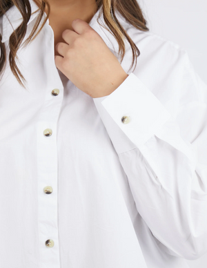 A person wearing a stylish Foxwood Paulina Shirt, made from 100% cotton poplin, adjusts the collar with their left hand. This white button-up design features gold buttons and long sleeves, set against a plain white background.