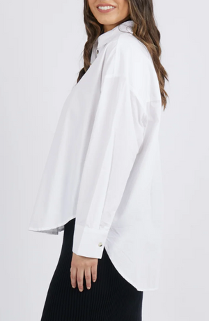 Side view of someone in a Foxwood Paulina Shirt, a 100% cotton poplin button-up with long sleeves, paired with a black skirt. They have long, wavy brown hair and are smiling against a crisp white background.