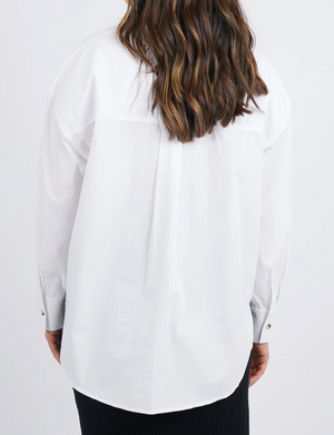 The image features a person with wavy brown hair wearing a Foxwood Paulina Shirt, a loose, stylish white long-sleeve made of 100% cotton poplin with buttoned cuffs. The relaxed-fit shirt is paired with black skirt or pants, against a plain white background, viewed from the back.