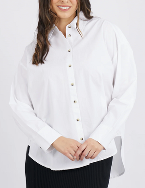 A person wearing a stylish Foxwood Paulina Shirt, made from 100% cotton poplin, and dark pants stands against a white background. The slightly oversized shirt has long sleeves and buttons down the front. The individual with long, wavy brown hair is smiling.