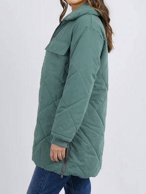 Against a light gray background, a person stands sideways wearing the Foxwood Whitley Jacketβa long, quilted green coat with zippered side splits and a large chest pocketβperfectly matched with blue jeans.