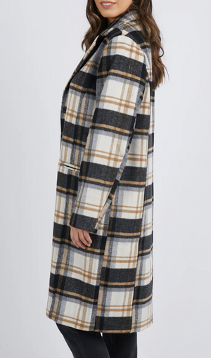 Wearing a Foxwood Catriona Coat in a chic plaid with black, white, and tan stripes over black pants, the person with loose wavy brown hair faces slightly sideways against a plain white backdrop. The coat’s collared design is crafted from polyester wool.