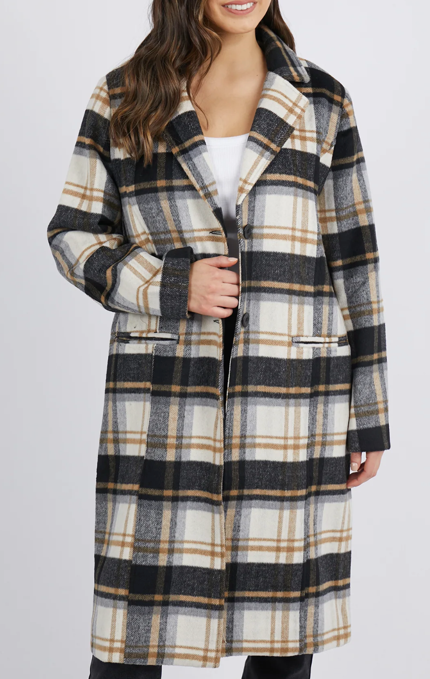 A person models the Foxwood Catriona Coat, a long plaid coat with a black, white, and brown checkered pattern. The polyester wool blend features a button closure, two pockets, and a collared design over a simple white top.