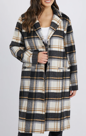 A person models the Foxwood Catriona Coat, a long plaid coat with a black, white, and brown checkered pattern. The polyester wool blend features a button closure, two pockets, and a collared design over a simple white top.