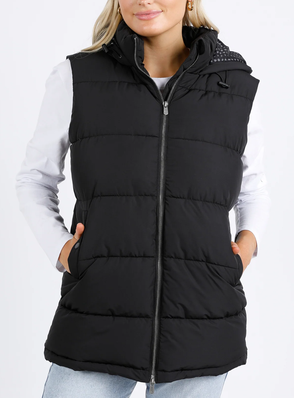 A person is wearing the cozy Foxwood Camilla Puffer Vest with an adjustable hood over a white long-sleeve shirt and light blue jeans, standing against a plain white background. Their hands rest casually in the vest pockets, enhancing its quilted detailing.