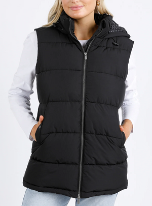 A person is wearing the cozy Foxwood Camilla Puffer Vest with an adjustable hood over a white long-sleeve shirt and light blue jeans, standing against a plain white background. Their hands rest casually in the vest pockets, enhancing its quilted detailing.