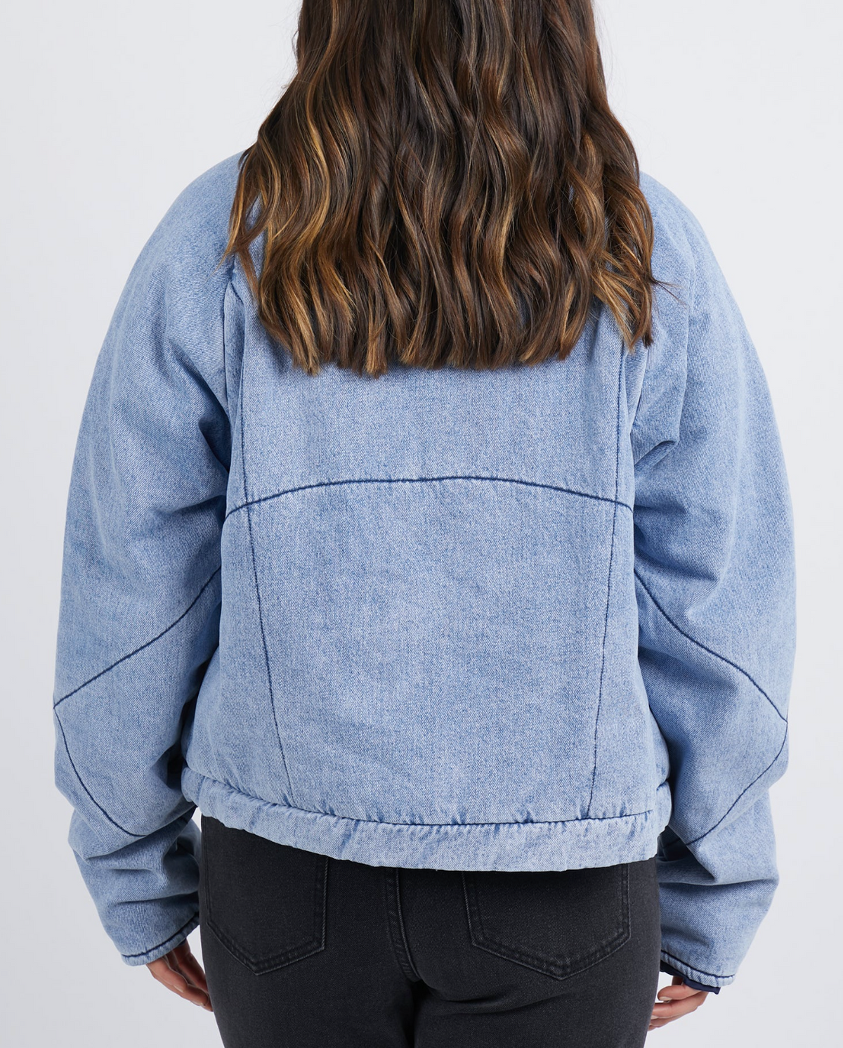 A person with wavy brown hair is shown from behind against a plain white background wearing the Foxwood Rosalee Denim Jacket, a light blue cropped cotton denim style, paired with black jeans.